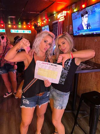 Two women in denim shorts pose inside a lively bar with colorful string lights and an American flag; they hold a printed sheet and make playful thumbs-down faces while one sticks her tongue out and a TV plays sports in the background.
