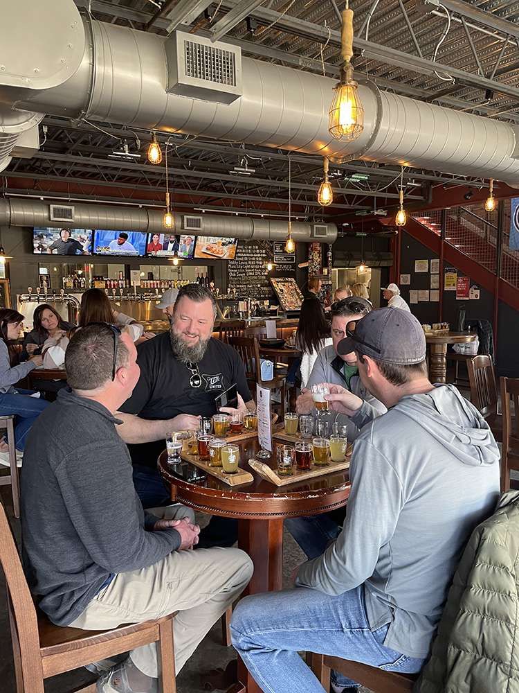 Four friends enjoying beer flights on wooden tasting paddles at a lively craft brewery taproom with exposed ductwork and hanging Edison bulbs.