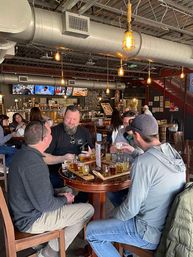 Four friends enjoying beer flights on wooden tasting paddles at a lively craft brewery taproom with exposed ductwork and hanging Edison bulbs.