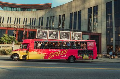 Bright pink-and-yellow open-top party bus with people waving on a sunlit downtown street in front of a large performing-arts building with black-and-white concert posters.