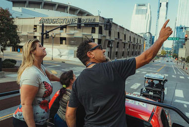 Tour guide pointing excitedly from an open-top red sightseeing vehicle while two passengers look up, passing a large arena and downtown skyscrapers on a city street.