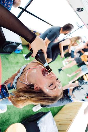 Young woman with glitter face jewels drinking from a bottle held by a friend at an outdoor daytime summer party on artificial turf, people dancing in the blurred background.