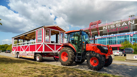 Orange tractor towing a red-and-white barn-style party wagon parked near a large stadium in a parking lot under a partly cloudy sky