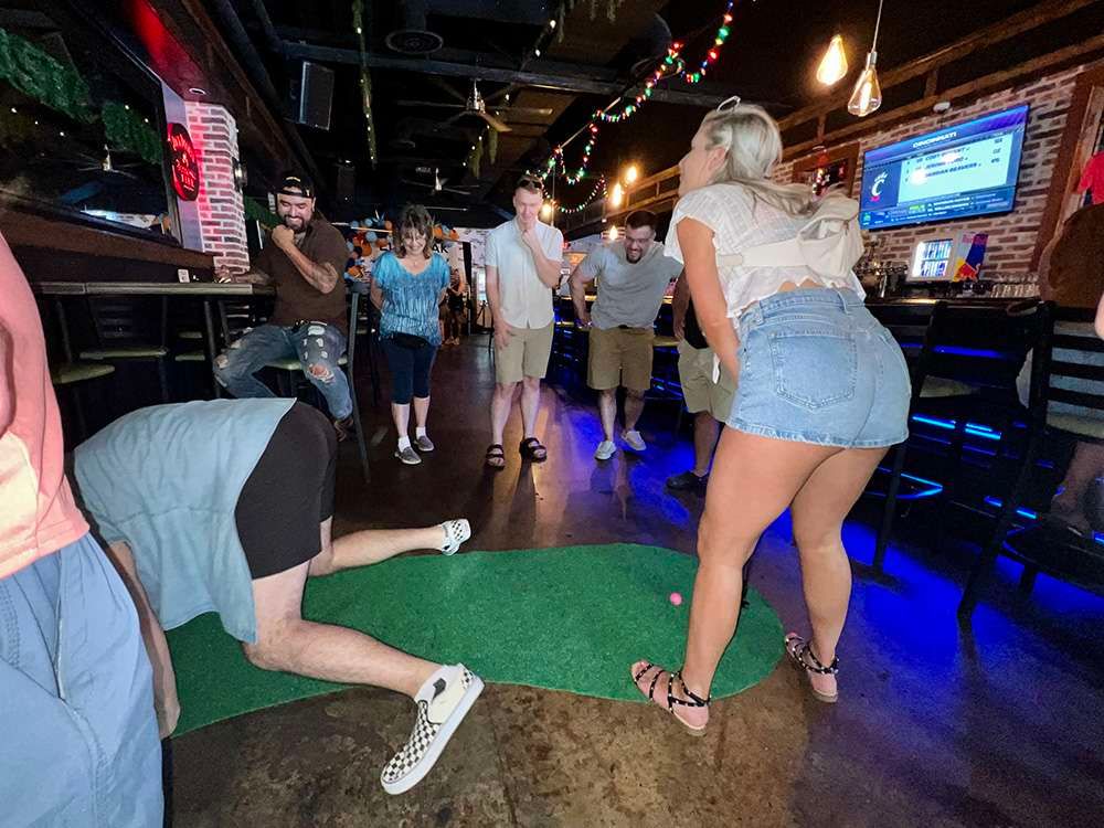 Group of friends playing indoor mini golf at a lively bar — woman in a denim skirt lining up a putt on a small green while another player crawls to retrieve the ball and others watch under string lights and neon bar lighting.