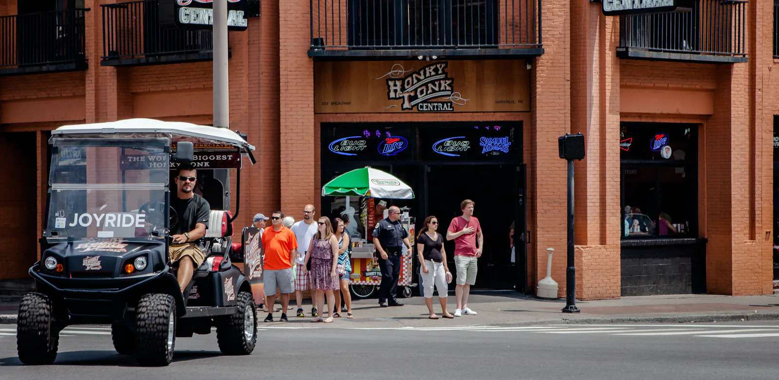 Lively downtown entertainment district street scene with an open-air tour vehicle passing a brick bar with balconies and neon signs; a group of pedestrians and a police officer wait by a sidewalk vendor under a green umbrella on a sunny day.