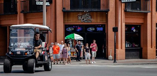 Lively downtown entertainment district street scene with an open-air tour vehicle passing a brick bar with balconies and neon signs; a group of pedestrians and a police officer wait by a sidewalk vendor under a green umbrella on a sunny day.