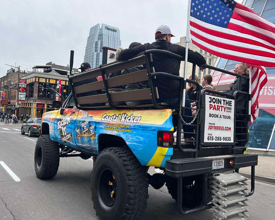 Colorful lifted party truck with bench seating and American flag carrying passengers down a downtown entertainment street lined with bars and high‑rise buildings.