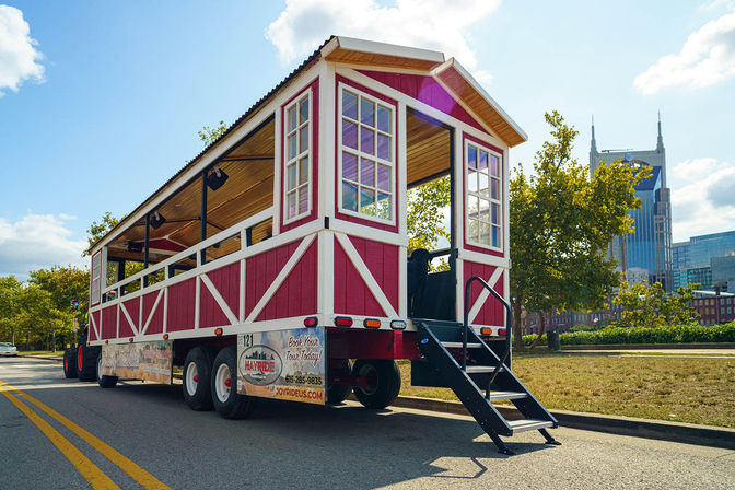 Red-and-white open-air hayride wagon parked on a sunny downtown Nashville street, black metal steps down to the pavement, leafy trees and a blue-sky city skyline in the background.