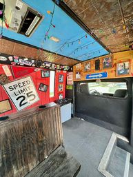 Quirky converted bus interior with wooden bench, Speed Limit 25 sign, framed photos, mini-fridge, tin ceiling and colorful string lights, retro roadside decor