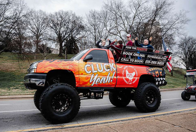 Orange chicken-themed lifted pickup truck with flame graphics and oversized off-road tires, passengers cheering in the open bed and an American flag, driving in a roadside parade on an overcast day with bare trees