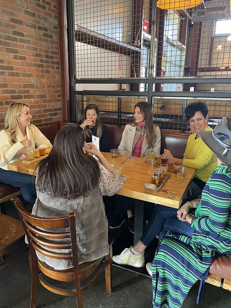 Six women laughing and sampling a craft beer flight at a wooden table in an industrial-style taproom with exposed brick and metal grid partition.