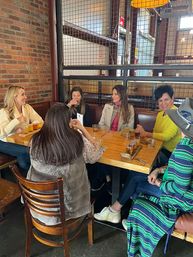 Six women laughing and sampling a craft beer flight at a wooden table in an industrial-style taproom with exposed brick and metal grid partition.