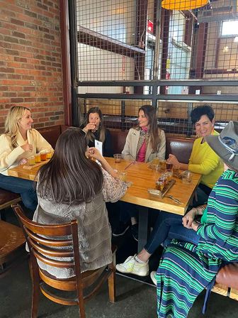 Six women laughing and sampling a craft beer flight at a wooden table in an industrial-style taproom with exposed brick and metal grid partition.