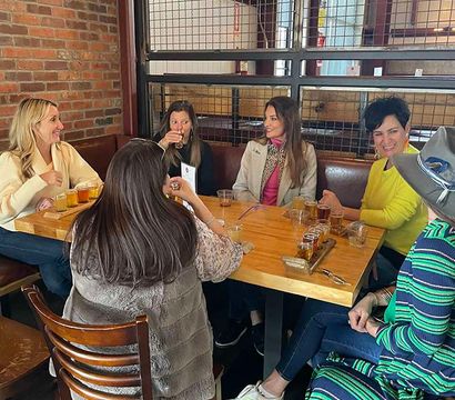 Six women laughing and sampling a craft beer flight at a wooden table in an industrial-style taproom with exposed brick and metal grid partition.