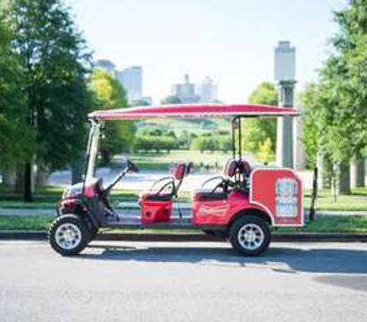 Red open-sided sightseeing cart parked by a tree-lined urban park with a distant downtown skyline on a sunny day