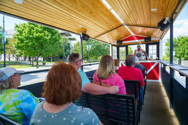 Open-air wood-roofed trolley tour with adults seated on bench seats listening to a standing guide at the red-and-white front, passing a tree-lined downtown street on a sunny day.
