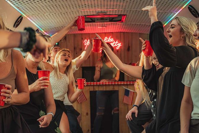 Lively group raising red solo cups inside a neon-lit party bus interior with wooden bar and small American flags.