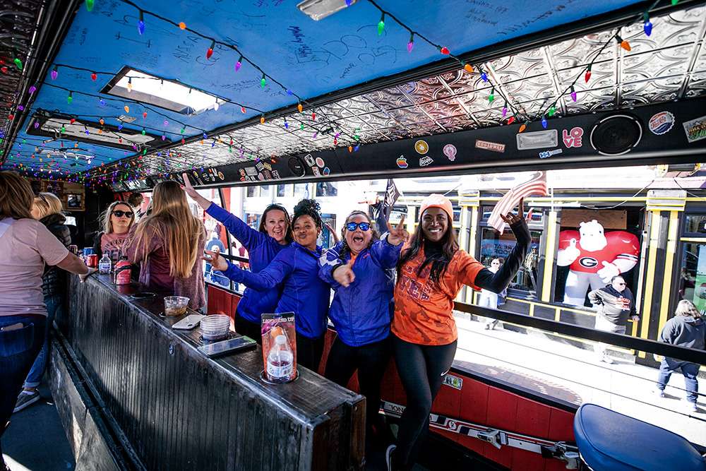 Group of people cheering inside a colorful open-sided party trolley decorated with string lights and stickers, overlooking a busy downtown street with a large inflatable mascot in a red jersey.