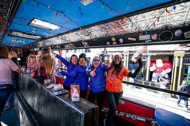 Group of people cheering inside a colorful open-sided party trolley decorated with string lights and stickers, overlooking a busy downtown street with a large inflatable mascot in a red jersey.