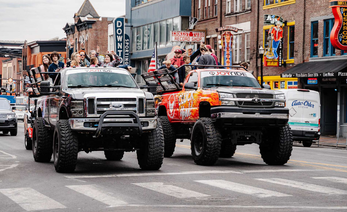 Two lifted pickup trucks with oversized tires side-by-side in a lively downtown parade, people standing and waving from open truck beds amid music-themed storefronts and an American flag.