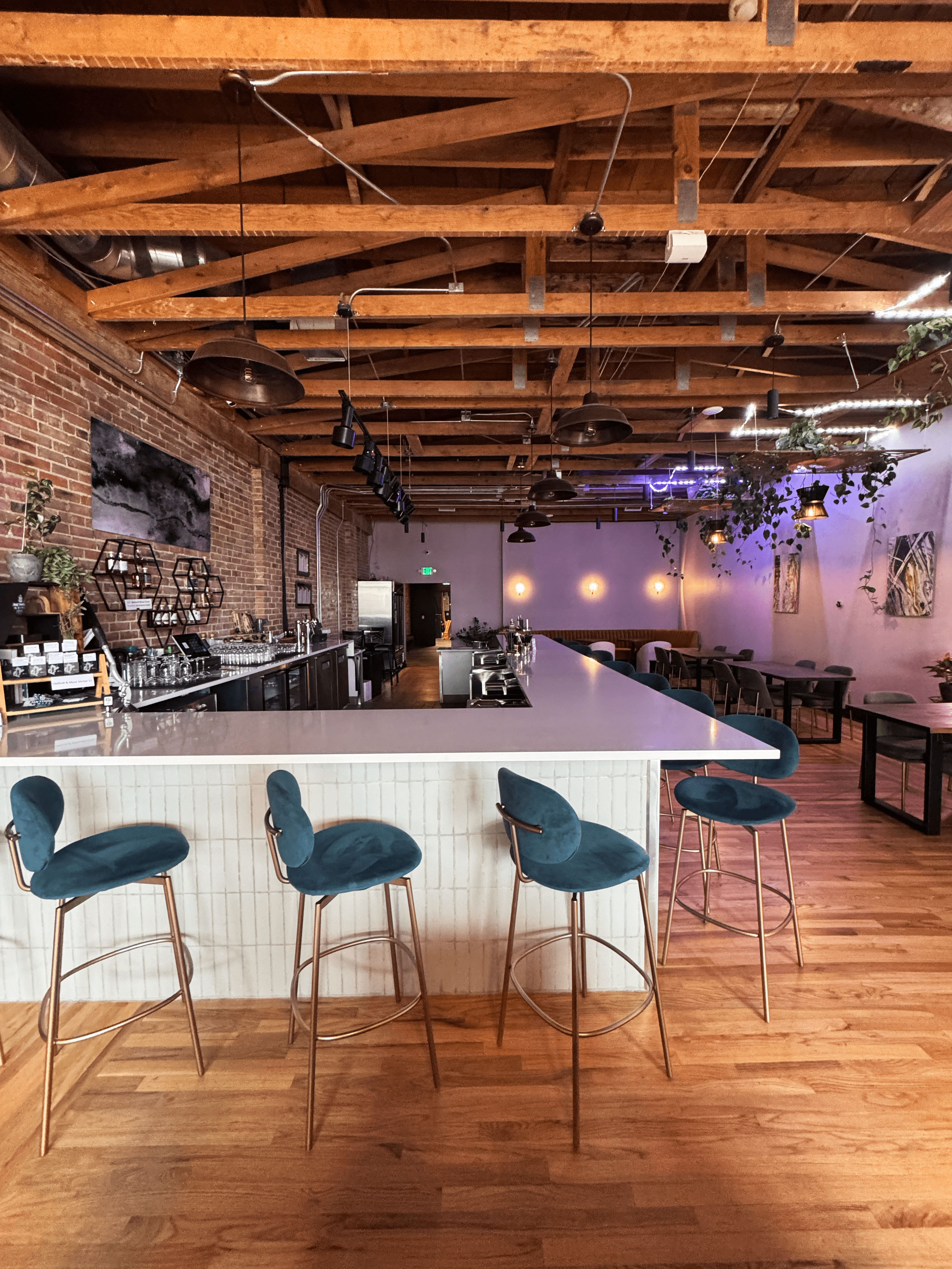 Loft-style café bar interior with a long white counter, teal velvet bar stools with gold legs, exposed wooden ceiling beams, brick accent wall, pendant lights, hanging plants and warm wooden floors.