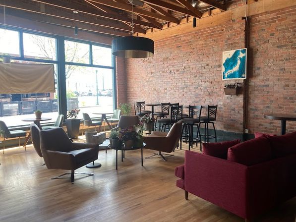 Sunlit urban café lounge with exposed brick wall, wooden floors, large windows, mixed seating of leather armchairs, high stools and a red sofa, and potted plants.