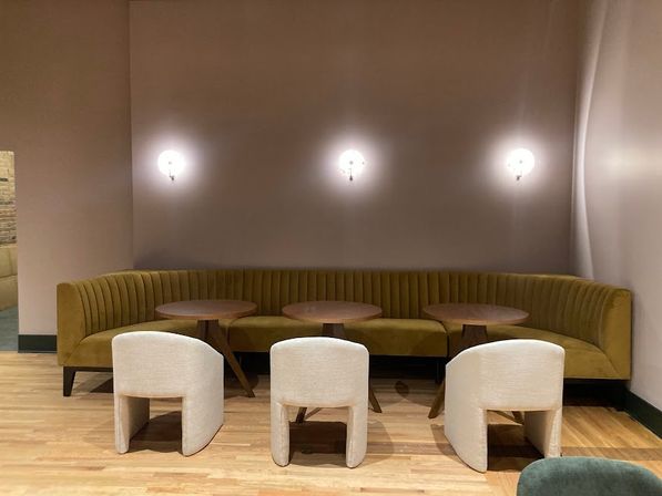 Modern indoor lounge with curved mustard-yellow banquette, three round wooden tables and three low cream upholstered chairs on light hardwood floor beneath soft wall sconces.