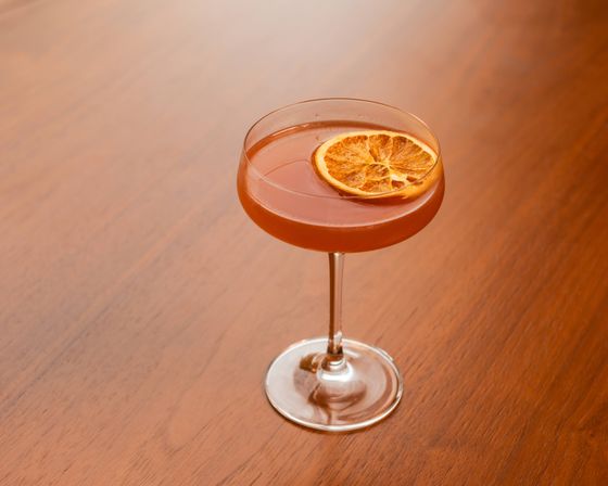 Amber craft cocktail in a coupe glass, garnished with a dried orange wheel, set on a warm wooden table.