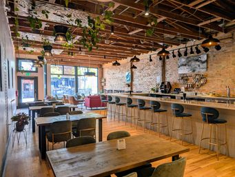 Sunlit industrial-chic downtown café interior with a long wooden bar and teal velvet barstools, communal wood tables, exposed brick wall, hanging plants and pendant lights by large front windows.