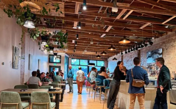 Vibrant urban cafe interior with exposed wooden beams, hanging plants and pendant lights; patrons chat at a bar with teal stools and at tables along an exposed-brick wall and large windows.