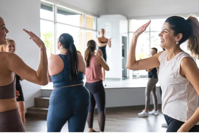 Women smiling and high-fiving during a group workout class in a bright indoor fitness studio with mirrored walls