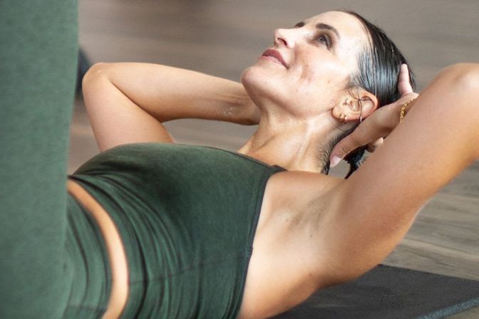Woman in green activewear doing abdominal crunches on a yoga mat indoors, hands behind her head and torso lifted, focused on core workout.