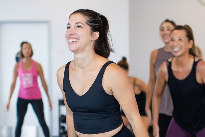 Smiling woman in a black workout top in the foreground of an energetic group fitness class in an indoor studio, other women exercising together.