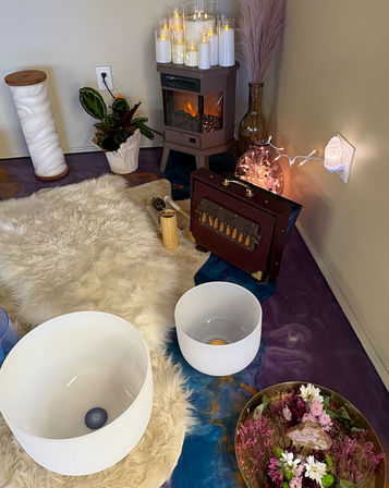 Cozy sound-bath meditation corner with two white crystal singing bowls on a fluffy sheepskin rug, small electric fireplace topped with LED candles, string lights, pampas grass, and a tray of flowers and crystals.