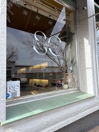 Downtown storefront window featuring a large white logo and hand-drawn floral vinyl decals, hanging globe lights and a potted plant inside, glass reflecting bare trees above a green-tiled window ledge.
