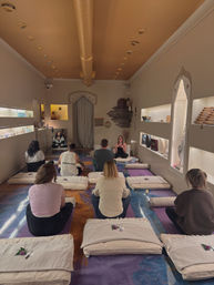 Group meditation and yoga class inside a cozy wellness studio with soft golden ceiling, recessed lighting, floor cushions and mats, participants seated facing an instructor amid decorative shelves and calming décor.