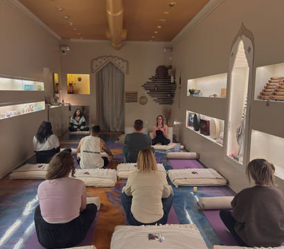 Group meditation and yoga class inside a cozy wellness studio with soft golden ceiling, recessed lighting, floor cushions and mats, participants seated facing an instructor amid decorative shelves and calming décor.