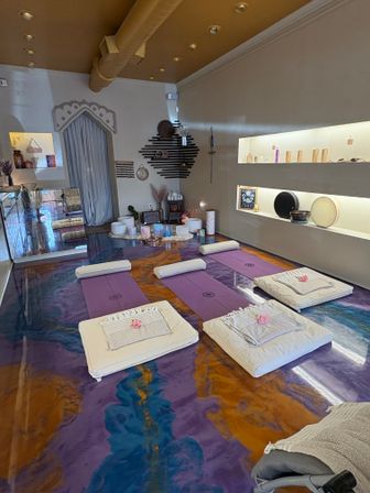 Calm wellness studio set for a sound bath: purple yoga mats and padded cushions topped with towels and pink roses, crystal singing bowls and instruments arranged on a central altar, marbled purple-and-gold epoxy floor, soft-lit shelves and decorative wall art.