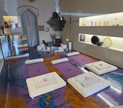 Calm wellness studio set for a sound bath: purple yoga mats and padded cushions topped with towels and pink roses, crystal singing bowls and instruments arranged on a central altar, marbled purple-and-gold epoxy floor, soft-lit shelves and decorative wall art.