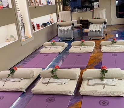 Meditation-ready yoga studio with rows of purple yoga mats, beige bolsters and folded blankets each topped with a single red rose, reclining chairs and softly lit wellness display shelves.