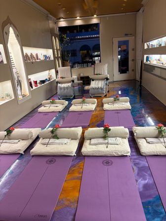 Boutique wellness studio interior with rows of purple yoga mats and cream bolsters, each topped with a single red rose, reclining chairs at the front, warm lit display shelves and a storefront window — set for a restorative yoga session.