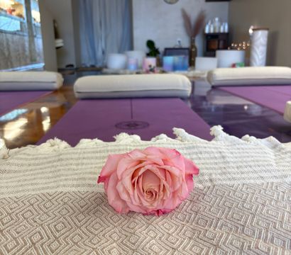 Pink rose on a textured blanket in the foreground with purple yoga mats, bolsters and soft lighting in a serene indoor yoga and meditation studio wellness space.