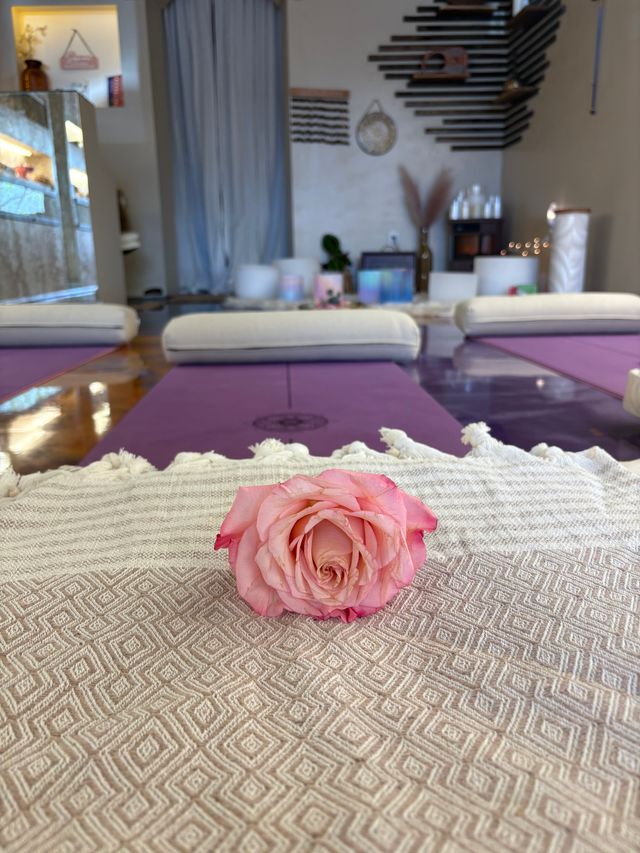 Pink rose on a textured blanket in the foreground with purple yoga mats, bolsters and soft lighting in a serene indoor yoga and meditation studio wellness space.