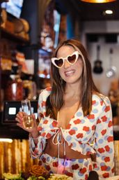 Smiling woman in a red-and-blue floral crop top and matching skirt, wearing glittery heart-shaped sunglasses and holding a champagne flute at a lively bar with plated food in the foreground