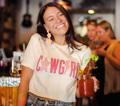 Smiling woman in a "Cowgirl" tee and studded denim shorts holding a bacon‑and‑olive garnished Bloody Mary in a cozy craft cocktail bar.