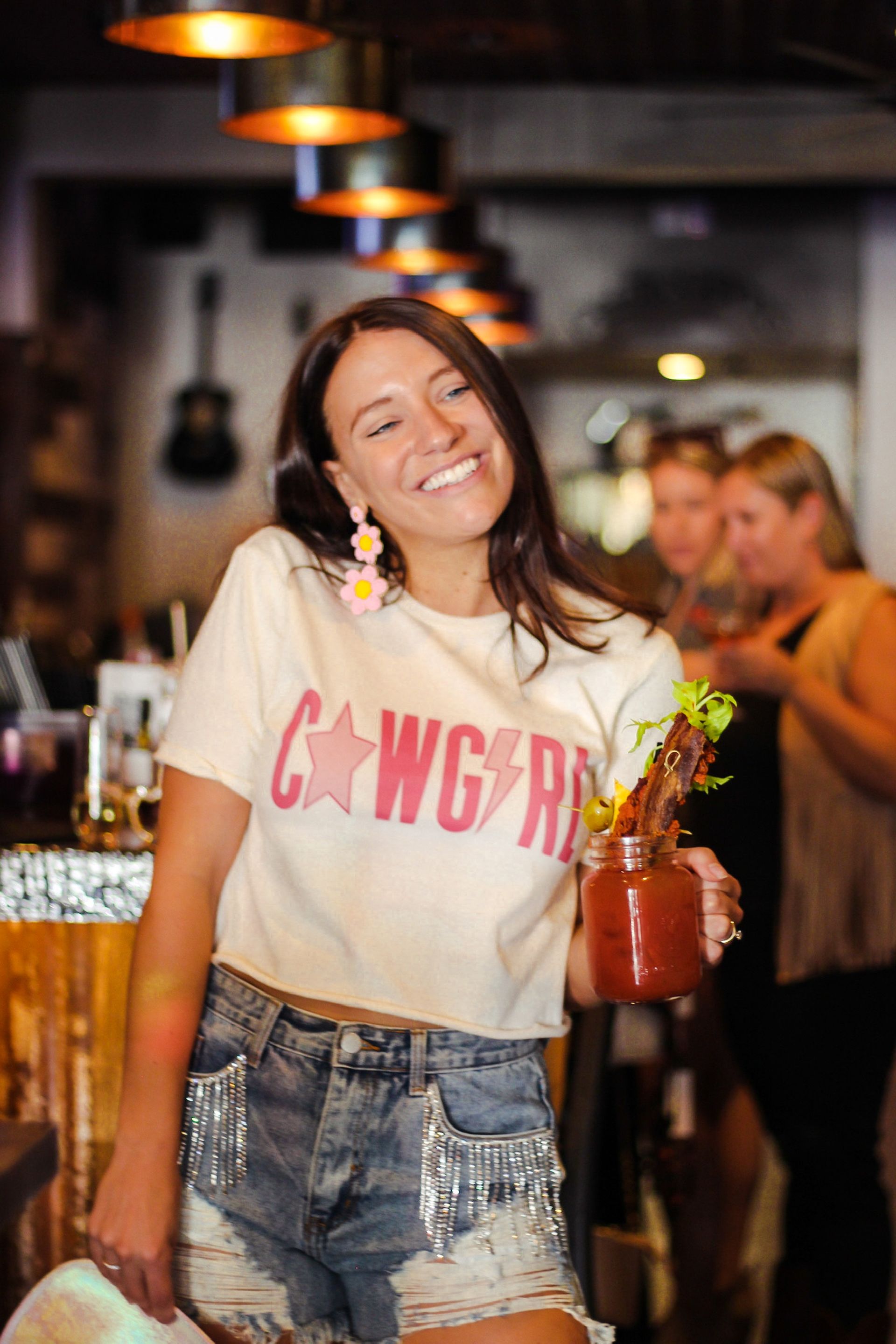 Smiling woman in a "Cowgirl" tee and studded denim shorts holding a bacon‑and‑olive garnished Bloody Mary in a cozy craft cocktail bar.
