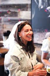 Smiling bride-to-be wearing a "BRIDE" veil and sparkly statement earrings, sipping a drink at a casual bachelorette brunch with rustic wood decor.