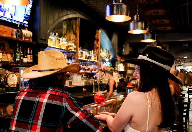Two people wearing cowboy hats laughing at a rustic saloon-style bar counter with a red cocktail, backlit liquor shelves and warm pendant lights.