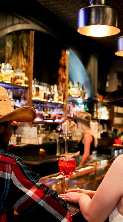 Smiling woman and man in cowboy hats chatting at a lively rustic western bar with illuminated liquor shelves and a red cocktail on the counter