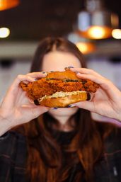 Close-up of a towering crispy fried chicken sandwich with coleslaw on a brioche bun, held up by a person in a casual restaurant under warm pendant lights.